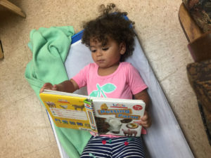 Toddler in childcare lying on a cot with a book curing naptime at daycare center